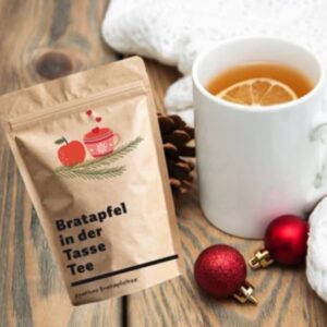 Bratapfeltee in brauner Tüte mit Teebecher und Weihnachtskugeln.